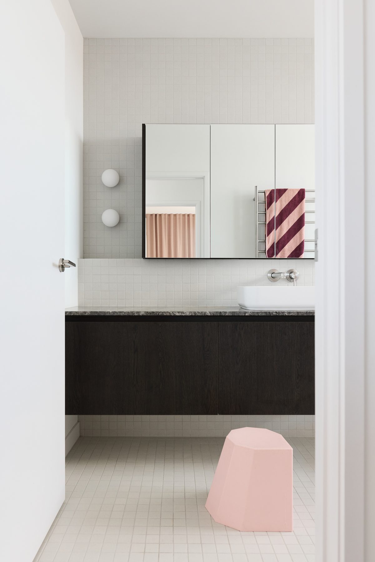 Bathroom in white mosaic, dark cabinetry and a pink stool.